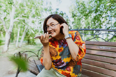 Happy woman sitting on bench in a green park, emotionally talking on her phone