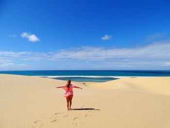 Full length rear view of woman with arms outstretched
on beach against blue sky