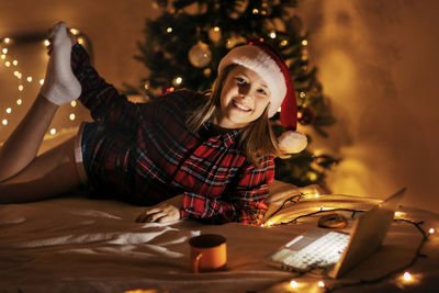 A teenage girl is lying on the bed next to a christmas tree decorated for christmas, using a laptop