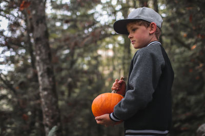 Side view of boy holding pumpkin against trees