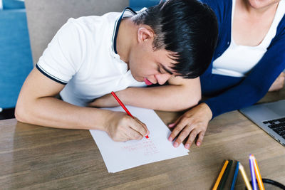 Ethnic teenage boy with down syndrome drawing with pencils on paper while sitting at table with cropped unrecognizable female freelancer working on laptop at home