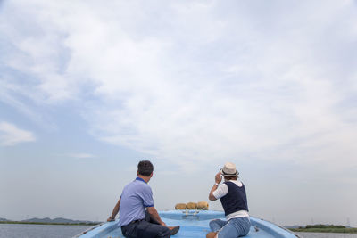 Rear view of men sitting on boat in river against cloudy sky