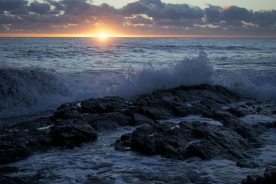 Scenic view of waves splashing on rocks against cloudy sky during sunset