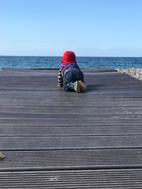 Rear view of boy crawling on boardwalk at beach against blue sky