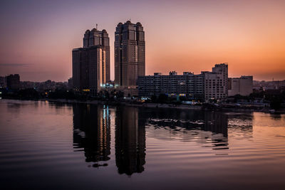 Illuminated buildings by lake against sky during sunset