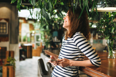 Young woman wearing striped longsleeve with mobile phone in hands in green eco cafe, city life