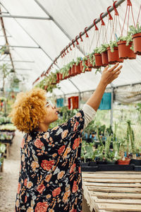 Side view of woman reaching for potted plants hanging on rod in nursery