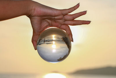 Close-up of hand holding crystal ball against sky during sunset