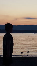 Silhouette boy standing by sea against sky during sunset