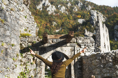 Rear view of woman in autumn clothes standing in old castle ruin, arms outstretched.
