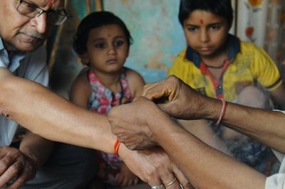 Cropped image of person tying thread to man hand while children looking at home