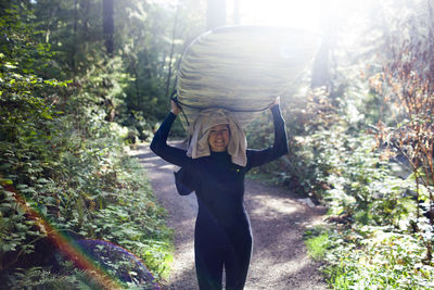 Happy woman carrying surfboard while standing on footpath amidst plants