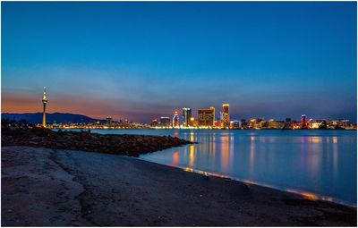 Illuminated city by sea against sky at sunset