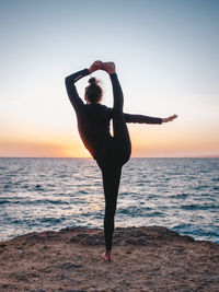 Full length of woman doing yoga at beach against sky during sunset