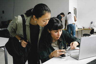 Customer standing near focused female technician examining laptop through magnifying glass at repair shop