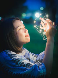 Young woman holding illuminated string lights while sitting on grassy field