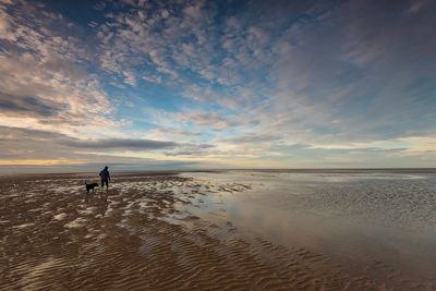 Rear view of woman walking with dog at beach against sky during sunset