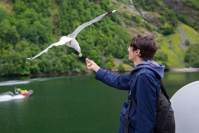 Young man feeding flying seagull while standing against lake