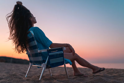 Woman sitting on chair at beach against sky during sunset