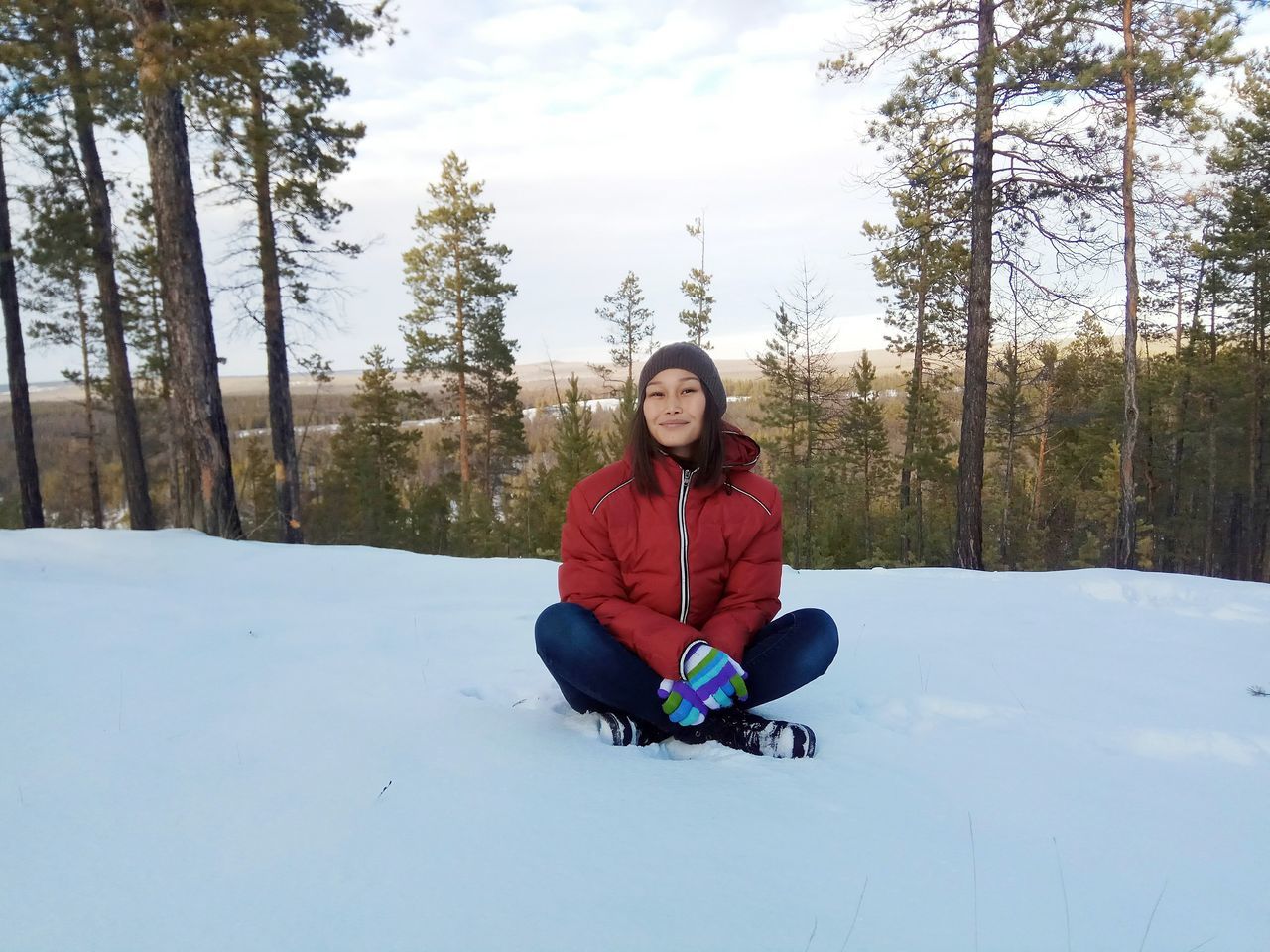 PORTRAIT OF SMILING WOMAN SITTING ON SNOW AGAINST TREES