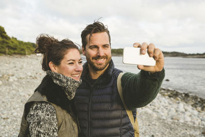 Happy couple taking selfie at beach against sky during sunset