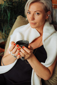 Stylish blond woman holding cup of coffee while sitting in cafe