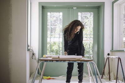 Female design professional talking on smart phone while examining blueprints on table at under construction apartment