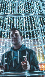 Low angle portrait of young man by illuminated string lights at night