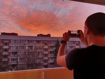 Rear view of man photographing cityscape against sky during sunset