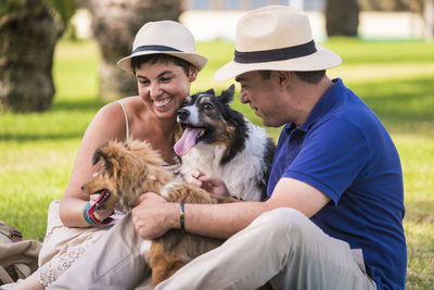 Smiling couple with dogs sitting on grassy field in park