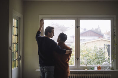 Rear view of romantic gay couple standing near window at home