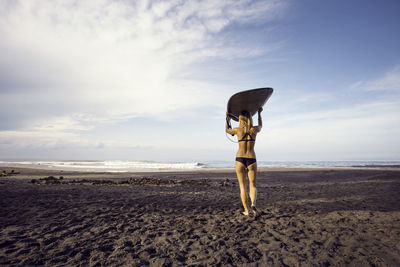 Rear view of woman carrying surfboard while walking at beach against sky