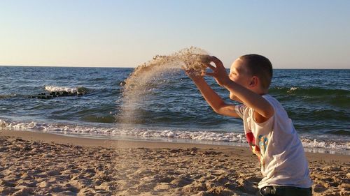Boy throwing sand at beach against clear sky