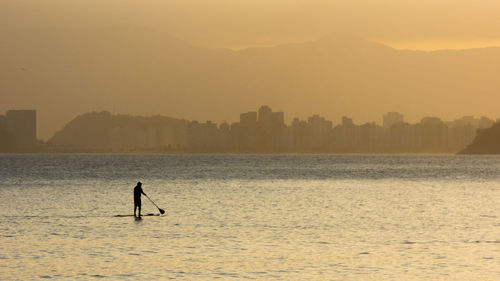 Silhouette person paddleboarding in river against city during sunset