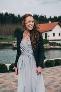 Smiling young woman standing on footpath by lake and house