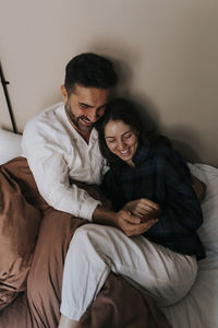 High angle view of happy couple sharing smart phone while sitting on bed at home