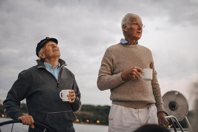 Male friends holding coffee cup while looking away against sky during sunset