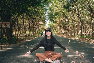 Portrait of smiling young woman throwing leaves while sitting on road amidst trees in forest