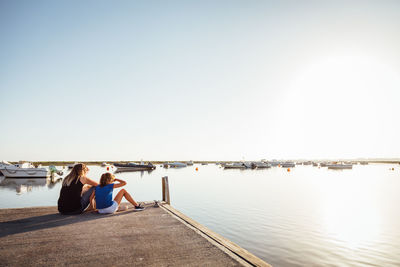 Rear view of mother and daughter sitting on pier at harbor against sky