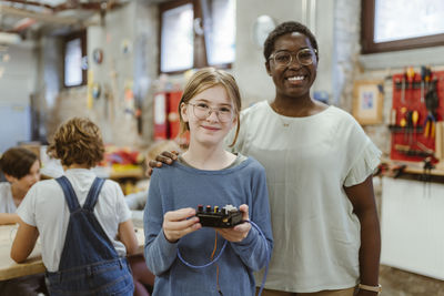 Portrait of smiling teacher with hand on shoulder of female student holding electrical part at technology workshop in sc