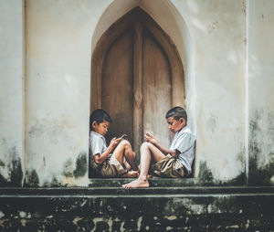 Side view of boys using phones while sitting at house entrance