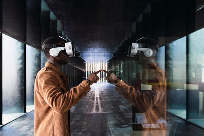 Side view of african american male in modern vr goggles standing touching a glass wall with reflection while exploring virtual reality
