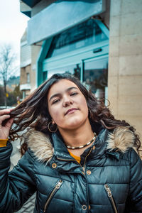 Portrait of young woman with hand in hair standing against building in city