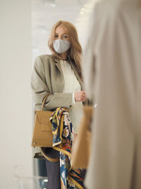 Young elegant woman with long blonde hair wearing face mask trying on a new classic bag in a mirror. 