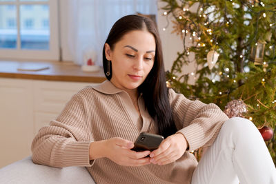 Woman with phone in hands sitting at home in kitchen on sofa during christmas person christmas tree