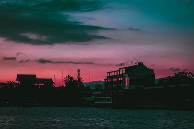 Silhouette buildings by sea against dramatic sky