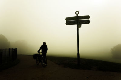 Man walking bicycle past crossroads sign  on road against foggy  sky