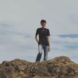 Low angle portrait of young woman standing on rock against sky