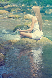 Side view of young woman meditating on rock in river
