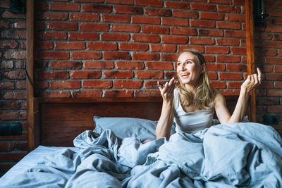 Adult woman with blonde long hair in pajamas using mobile phone sitting on bed in loft room at home